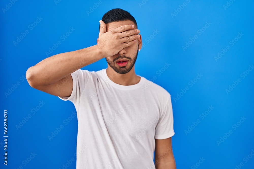 Poster Young hispanic man standing over blue background covering eyes with hand, looking serious and sad. sightless, hiding and rejection concept