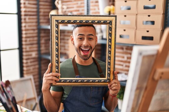 Young hispanic man sitting at art studio with empty frame celebrating crazy and amazed for success with open eyes screaming excited.