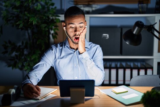 Young hispanic man working at the office at night afraid and shocked, surprise and amazed expression with hands on face