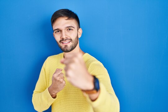 Hispanic man standing over blue background punching fist to fight, aggressive and angry attack, threat and violence