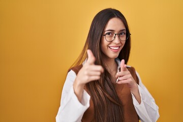 Young brunette woman standing over yellow background wearing glasses pointing fingers to camera with happy and funny face. good energy and vibes.