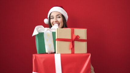 Beautiful hispanic woman wearing christmas hat holding gifts over isolated red background