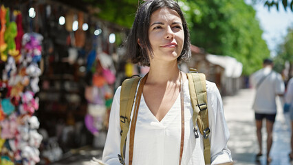 Young beautiful hispanic woman tourist wearing backpack standing at street market