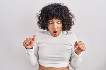 Hispanic woman with curly hair standing over isolated background pointing down with fingers showing advertisement, surprised face and open mouth