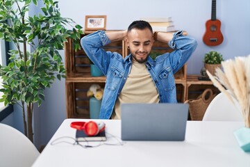 Young hispanic man sitting on table relaxed with hands on head at home