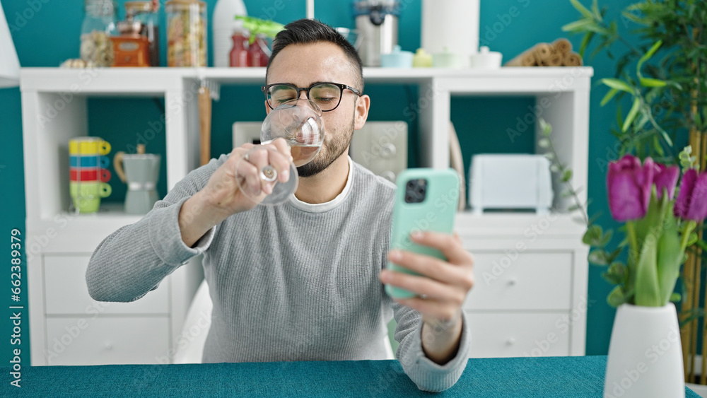 Wall mural Hispanic man drinking glass of wine sitting on table using smartphone at dinning room