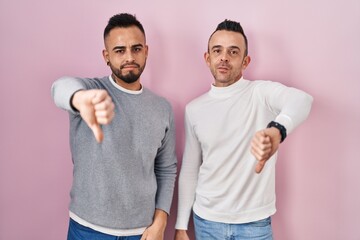 Homosexual couple standing over pink background looking unhappy and angry showing rejection and negative with thumbs down gesture. bad expression.