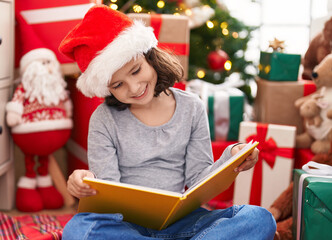 Fototapeta na wymiar Adorable chinese girl reading book sitting on floor by christmas tree at home