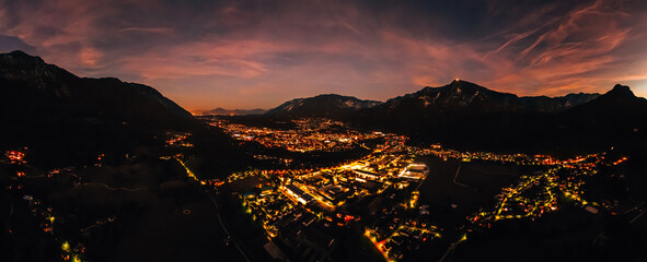 Aerial view of the lights in a small city in a valley of the Alps mountains at night as pink sunset clouds are in the sky