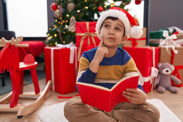 Obraz na płótnie Canvas Adorable hispanic boy reading book sitting on floor by christmas tree at home