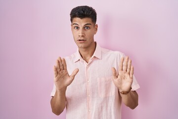 Young hispanic man standing over pink background moving away hands palms showing refusal and denial with afraid and disgusting expression. stop and forbidden.