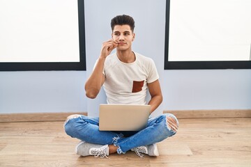 Young hispanic man using laptop at home mouth and lips shut as zip with fingers. secret and silent,...