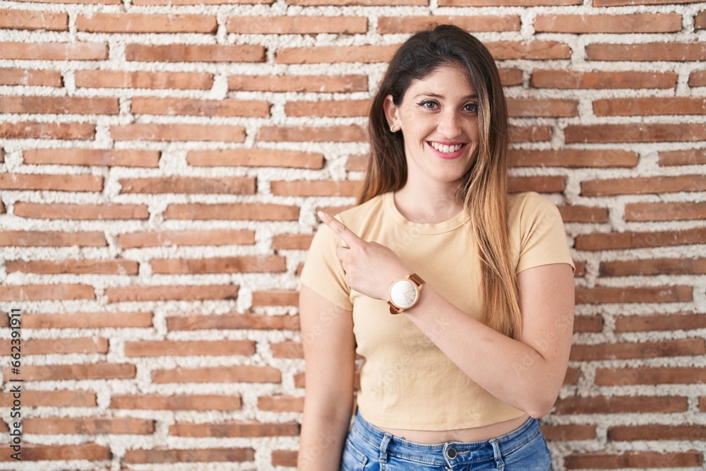 Poster Young brunette woman standing over bricks wall cheerful with a smile on face pointing with hand and finger up to the side with happy and natural expression