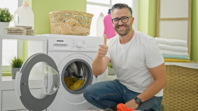Grey-haired man washing clothes doing thumb up gesture smiling at laundry room