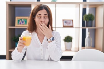 Brunette woman drinking glass of orange juice bored yawning tired covering mouth with hand. restless and sleepiness.