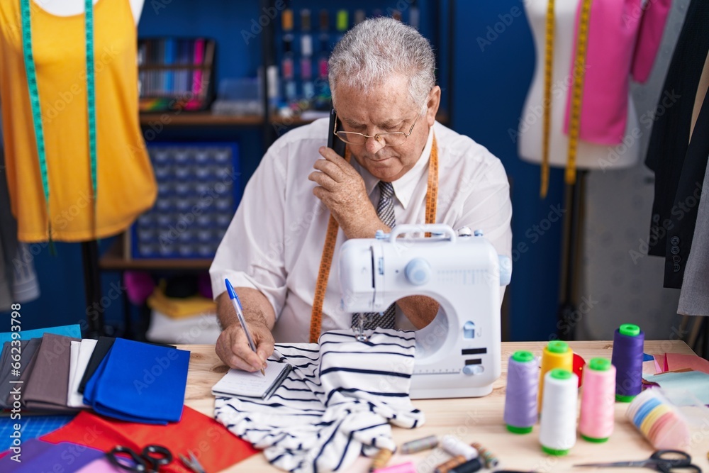 Canvas Prints Middle age grey-haired man tailor talking on smartphone writing on notebook at tailor shop