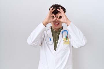 Young non binary man wearing doctor uniform and stethoscope doing ok gesture like binoculars sticking tongue out, eyes looking through fingers. crazy expression.