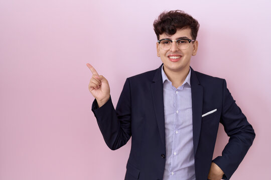 Young non binary man with beard wearing suit and tie with a big smile on face, pointing with hand and finger to the side looking at the camera.