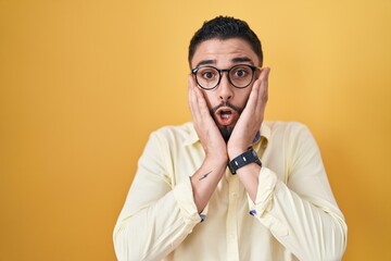 Hispanic young man wearing business clothes and glasses afraid and shocked, surprise and amazed expression with hands on face