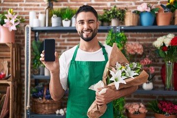 Hispanic young man working at florist shop showing smartphone screen winking looking at the camera...