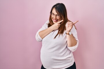 Pregnant woman standing over pink background pointing aside worried and nervous with both hands,...
