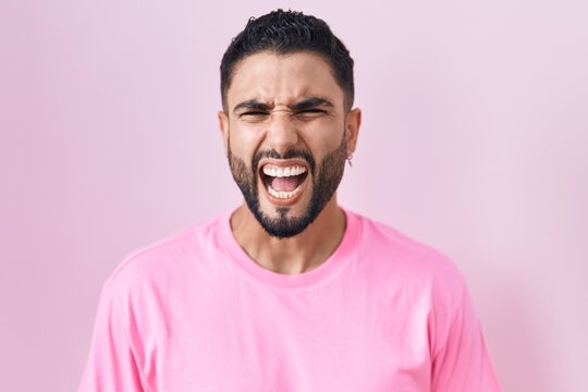 Hispanic young man standing over pink background angry and mad screaming frustrated and furious, shouting with anger. rage and aggressive concept.