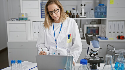 Naklejka premium Dedicated young blonde scientist concentrates on her laptop, engrossed in vital medical research in the bustling heart of a vibrant lab
