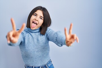 Young hispanic woman standing over blue background smiling with tongue out showing fingers of both...