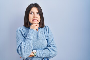 Young hispanic woman standing over blue background thinking worried about a question, concerned and nervous with hand on chin