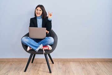 Young hispanic woman sitting on chair using computer laptop smiling with happy face winking at the camera doing victory sign with fingers. number two. - obrazy, fototapety, plakaty
