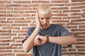Young caucasian man standing over bricks wall looking at the watch time worried, afraid of getting...