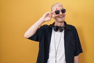 Young caucasian man wearing sunglasses standing over yellow background doing peace symbol with fingers over face, smiling cheerful showing victory