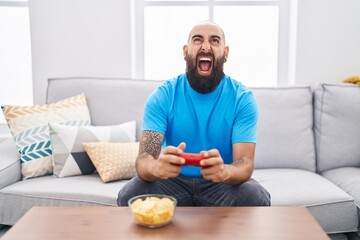 Young hispanic man with beard and tattoos playing video game sitting on the sofa angry and mad screaming frustrated and furious, shouting with anger looking up.