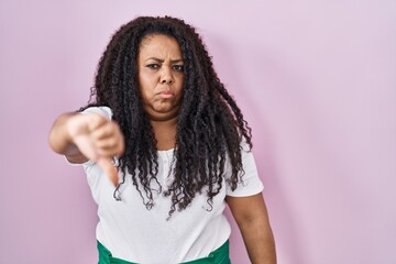 Plus size hispanic woman standing over pink background looking unhappy and angry showing rejection and negative with thumbs down gesture. bad expression.