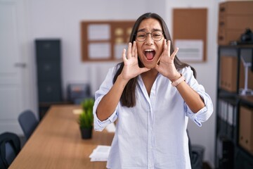 Young hispanic woman at the office shouting angry out loud with hands over mouth