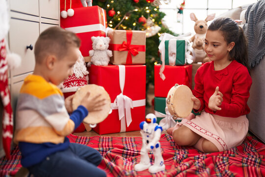 Brother and sister playing tambourine sitting on floor by christmas gifts at home