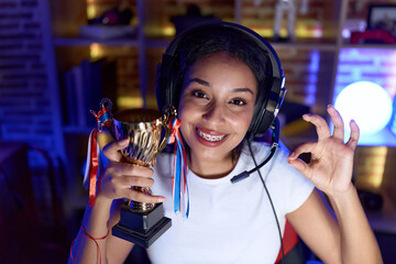 Young arab woman playing video games holding trophy doing ok sign with fingers, smiling friendly...