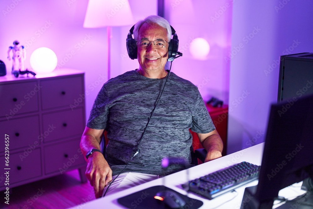 Poster middle age grey-haired man streamer smiling confident sitting on table at gaming room