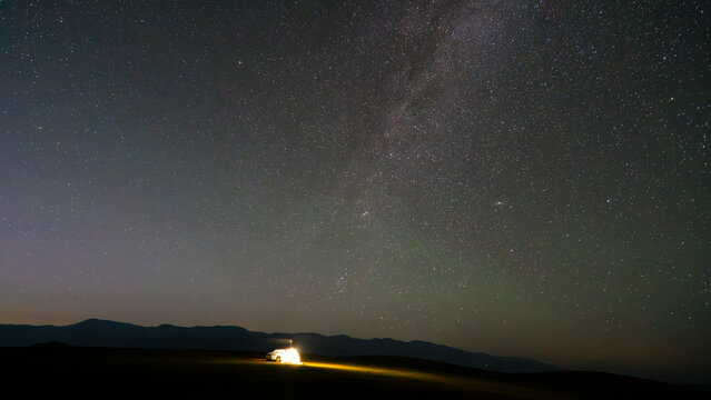 The starry sky at sunset. The tent and the car in the field are lit up. Hills are visible in the distance. Meteorites are falling. The Milky Way, many stars and the Andromeda galaxy. Photos on desktop