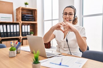 Young hispanic woman wearing call center agent headset at the office smiling in love showing heart...