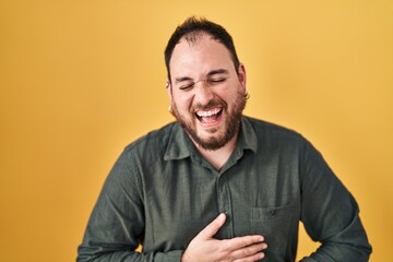 Plus size hispanic man with beard standing over yellow background smiling and laughing hard out loud because funny crazy joke with hands on body.