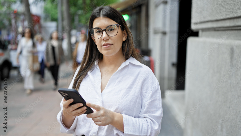 Wall mural young beautiful hispanic woman using smartphone with serious face in the streets of madrid