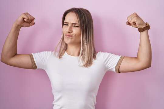 Blonde caucasian woman standing over pink background showing arms muscles smiling proud. fitness concept.