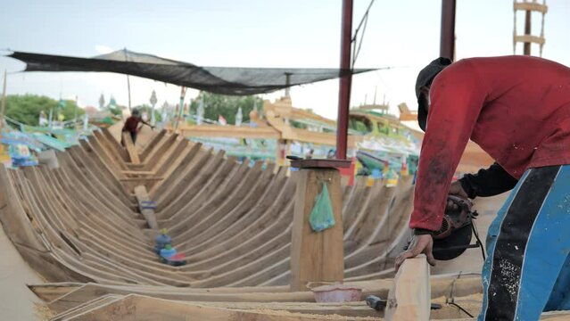A shipbuilder cuts the timber of a traditional ship's frame on the Indonesian coast with other workers working on the deck