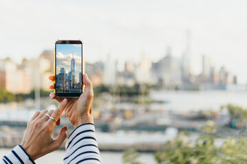 A person takes a mobile photo of the New York City's skyline seen from the bank of Hudson river at the West side of New York City, United States. - obrazy, fototapety, plakaty