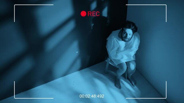 High angle CCTV shot of young man with schizophrenia wearing straitjacket sitting on chair in corner of ward grimacing, screaming and shaking head