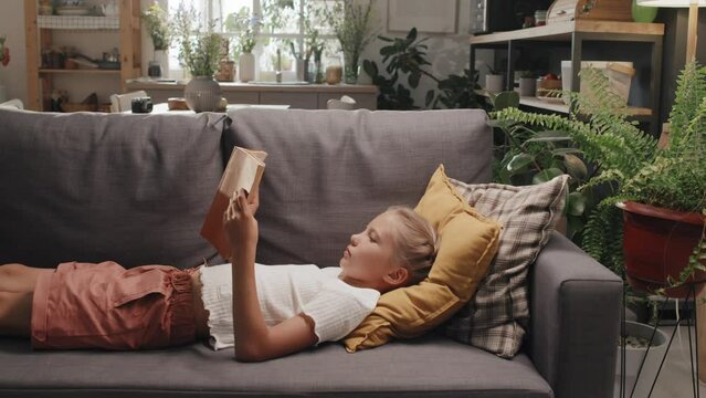 Medium long shot of preteen girl relaxing on couch in living room at home reading book