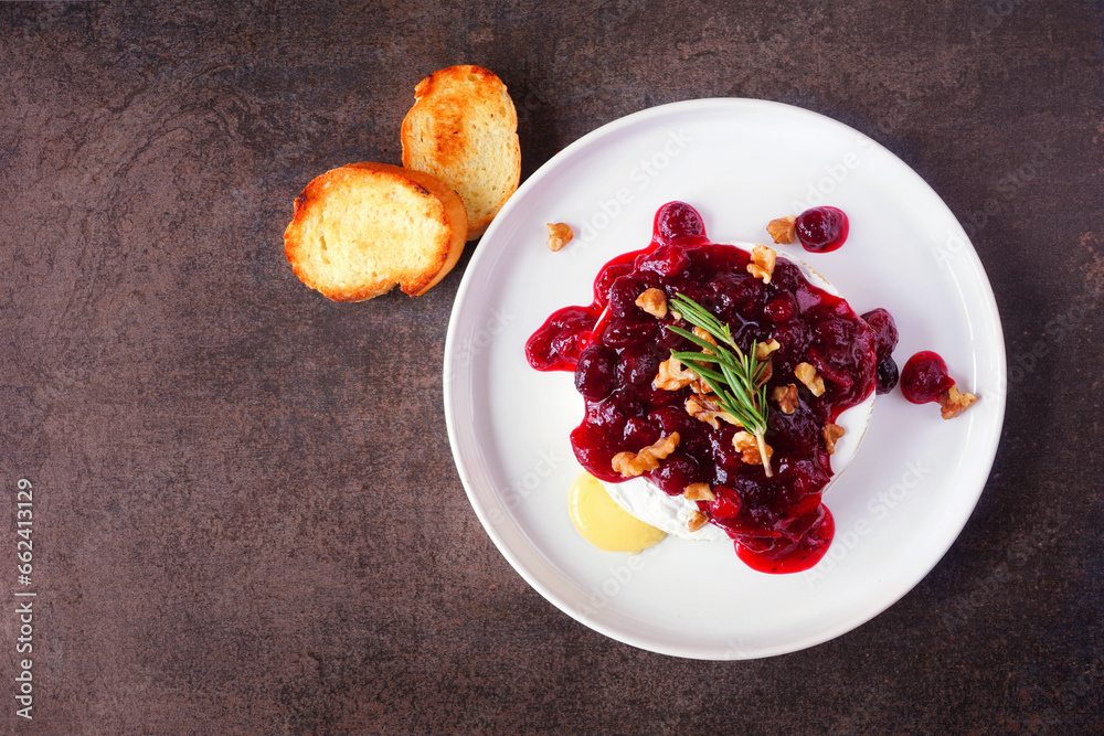 Poster baked brie appetizer garnished with cranberry sauce and walnuts. overhead view on a dark background.