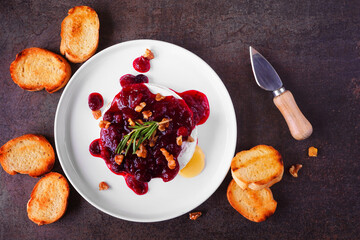 Baked brie appetizer garnished with cranberry sauce and walnuts. Top down view table scene with toasted baguette over a dark background.