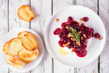 Baked brie appetizer garnished with cranberry sauce and walnuts. Above view table scene with toasted baguette over a white wood background.